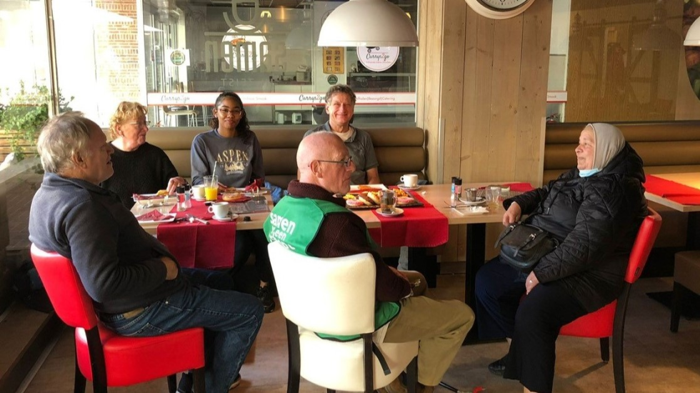 Zes vrijwilligers uit Vollenhove zitten aan tafel in Samen in Zeist, waar ze koffie drinken met iets lekkers. 