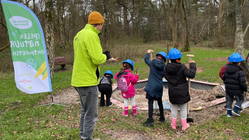 Een foto van 5 kinderen met een blauwe helm op en een schep in hun hand staan in het park. Er staat ook een begeleider een vlag van Gezonde Buurten bij. 