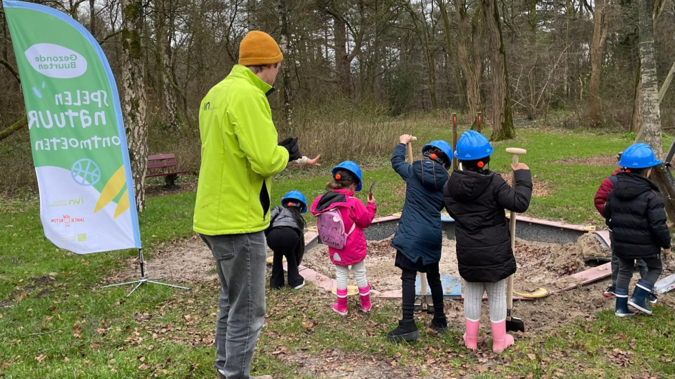 Een foto van 5 kinderen met een blauwe helm op en een schep in hun hand staan in het park. Er staat ook een begeleider een vlag van Gezonde Buurten bij.