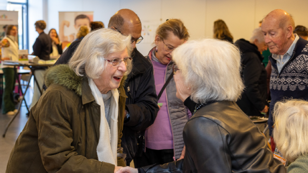 Twee vrouwen schudden elkaar de hand bij een kraam met flyers op het Gezondheidsfestival. Op de achtergrond lopen meerdere mensen