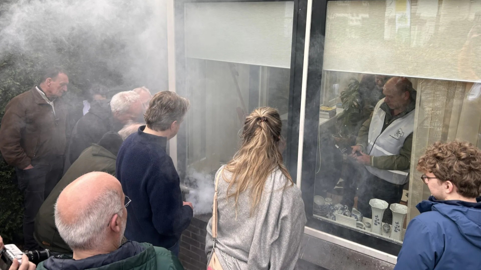 Een groep van zes bewoners kijken bij een kozijn naar de rook die eruit komt tijdens de blowerdoortest