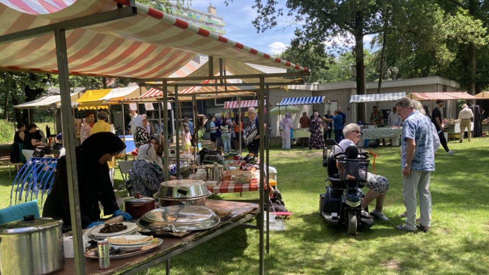 Foto van het Buurtfeest in het Bospark. Een kraampje met eten links en rechts twee bewoners die met elkaar praten.
